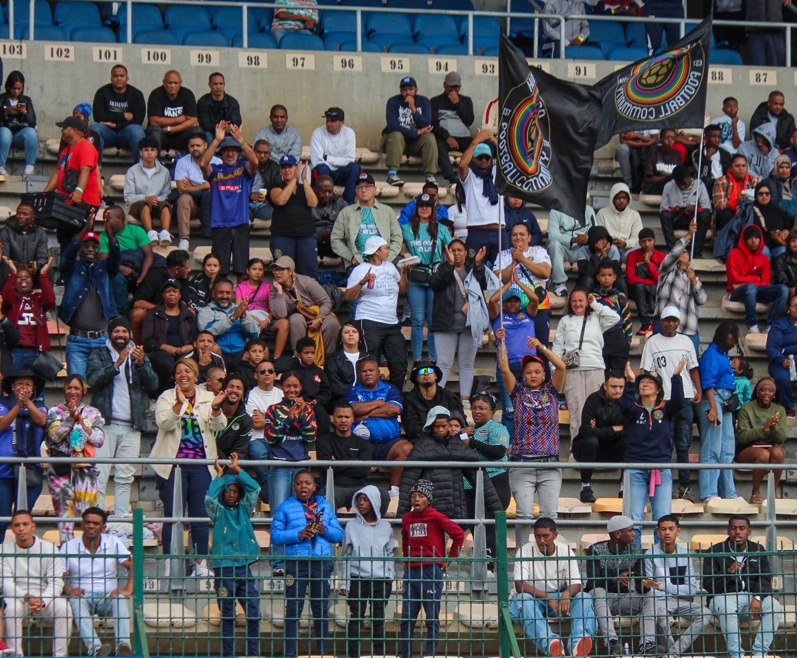 Hout Bay United supporters on their feet in the stands, clapping, with the club flag flying